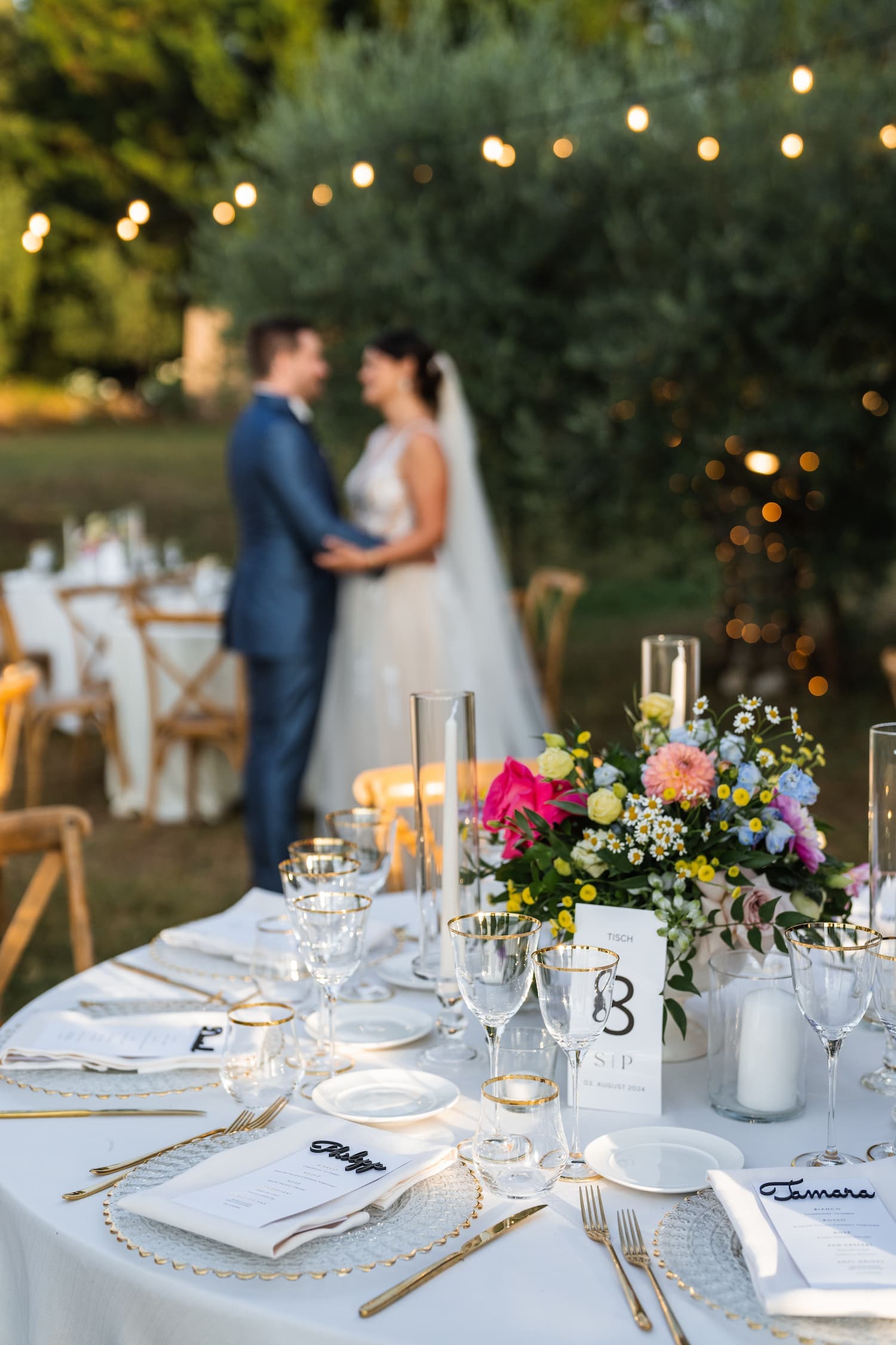 Liebevoll gedeckter Hochzeitstisch mit Wildblumen und Lichterketten, im Hintergrund ein Brautpaar