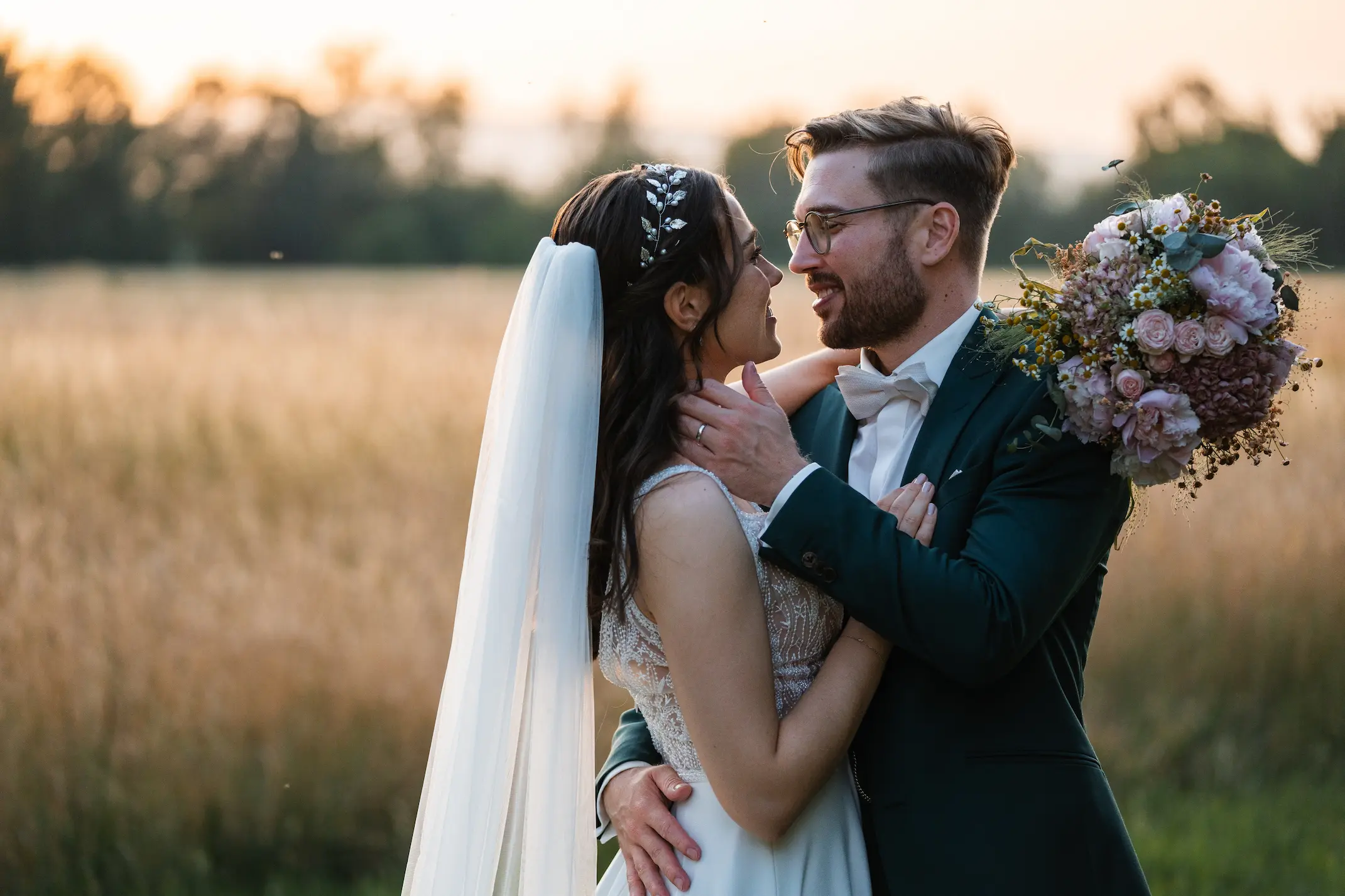 Brautpaar im Sonnenuntergang an einem Getreidefeld, die Braut mit Schleier und der Bräutigam mit Brautstrauß – Hochzeit organisiert mit celebrateUs